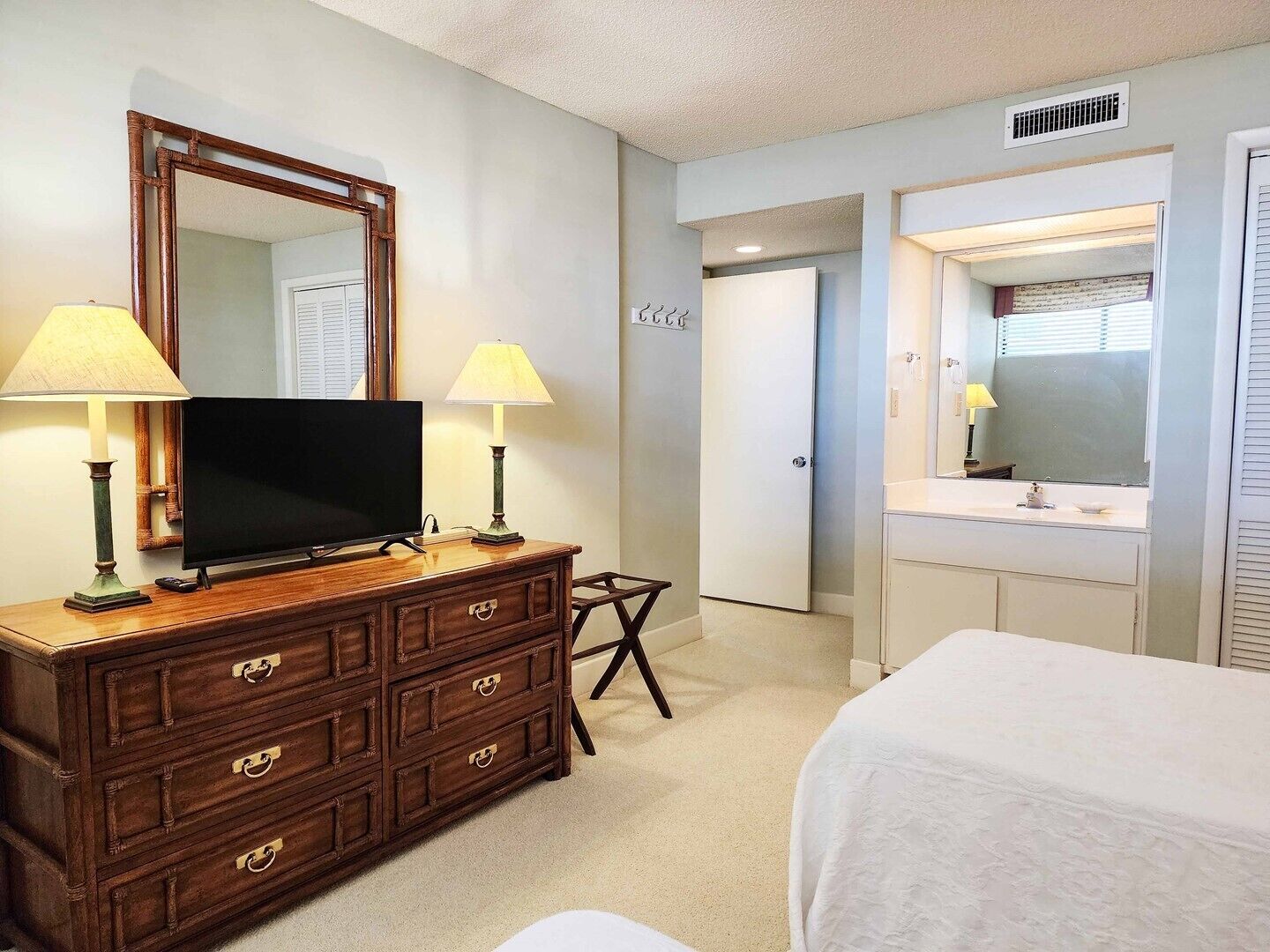 Une chambre d’hôtel avec une commode en bois, un téléviseur à écran plat, deux lampes de chevet, un lit, une salle de bain avec un miroir et une porte portant l’inscription « ÉCOLE ».