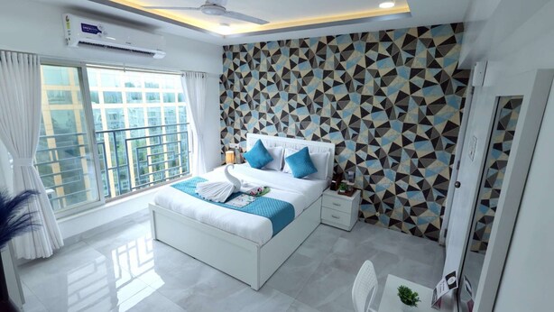 A modern bedroom with a bed, a ceiling fan, a window with curtains, and a geometric patterned wall. - 1 BHK Suite ( Gagal Home) (Mumbai)