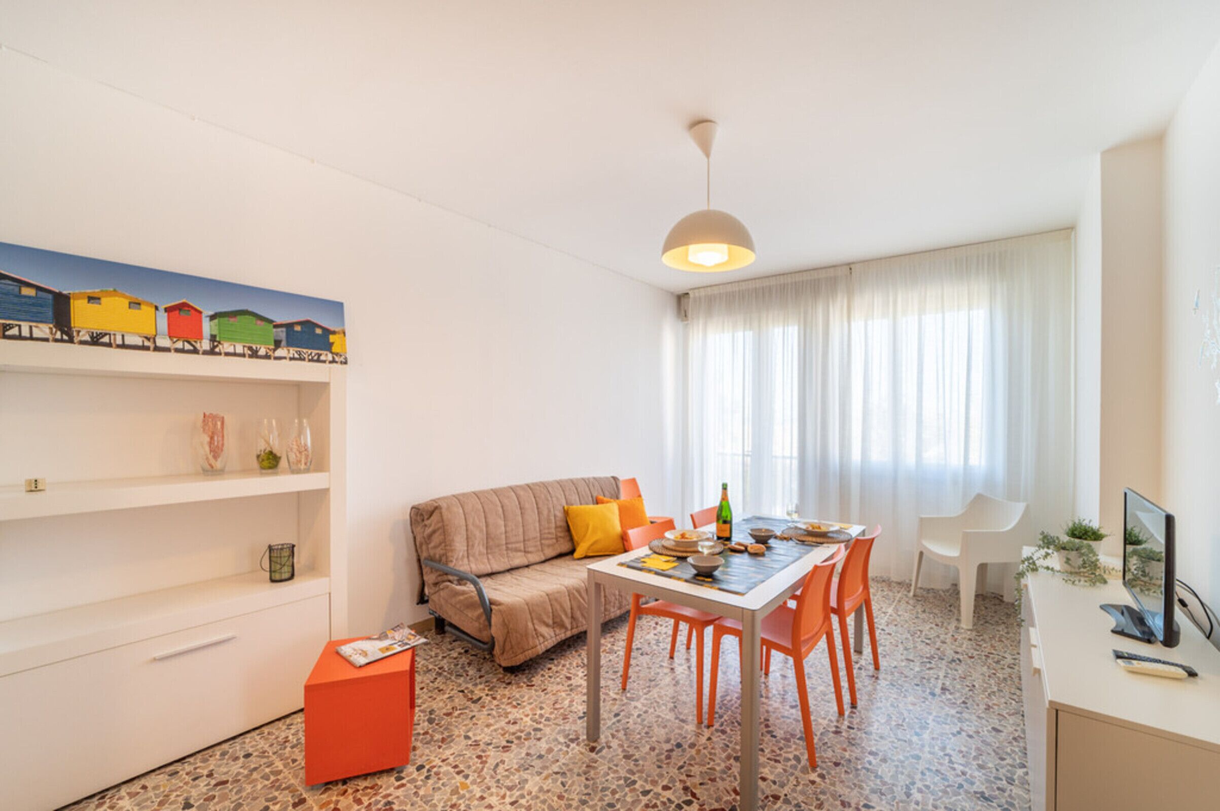 Un salon moderne avec un canapé, une table à manger et des chaises. Il y a un tableau au mur, un luminaire au plafond et une fenêtre avec des rideaux.