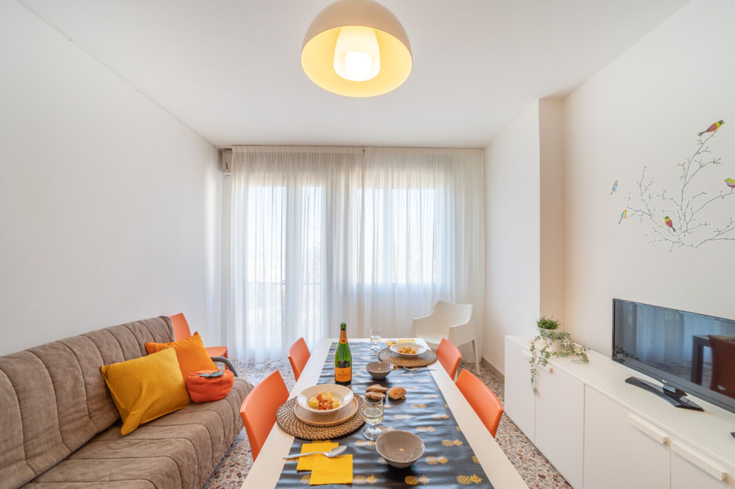 A modern living room with a dining table set for four, a sofa with orange cushions, a flat-screen TV, and a large window with sheer curtains.