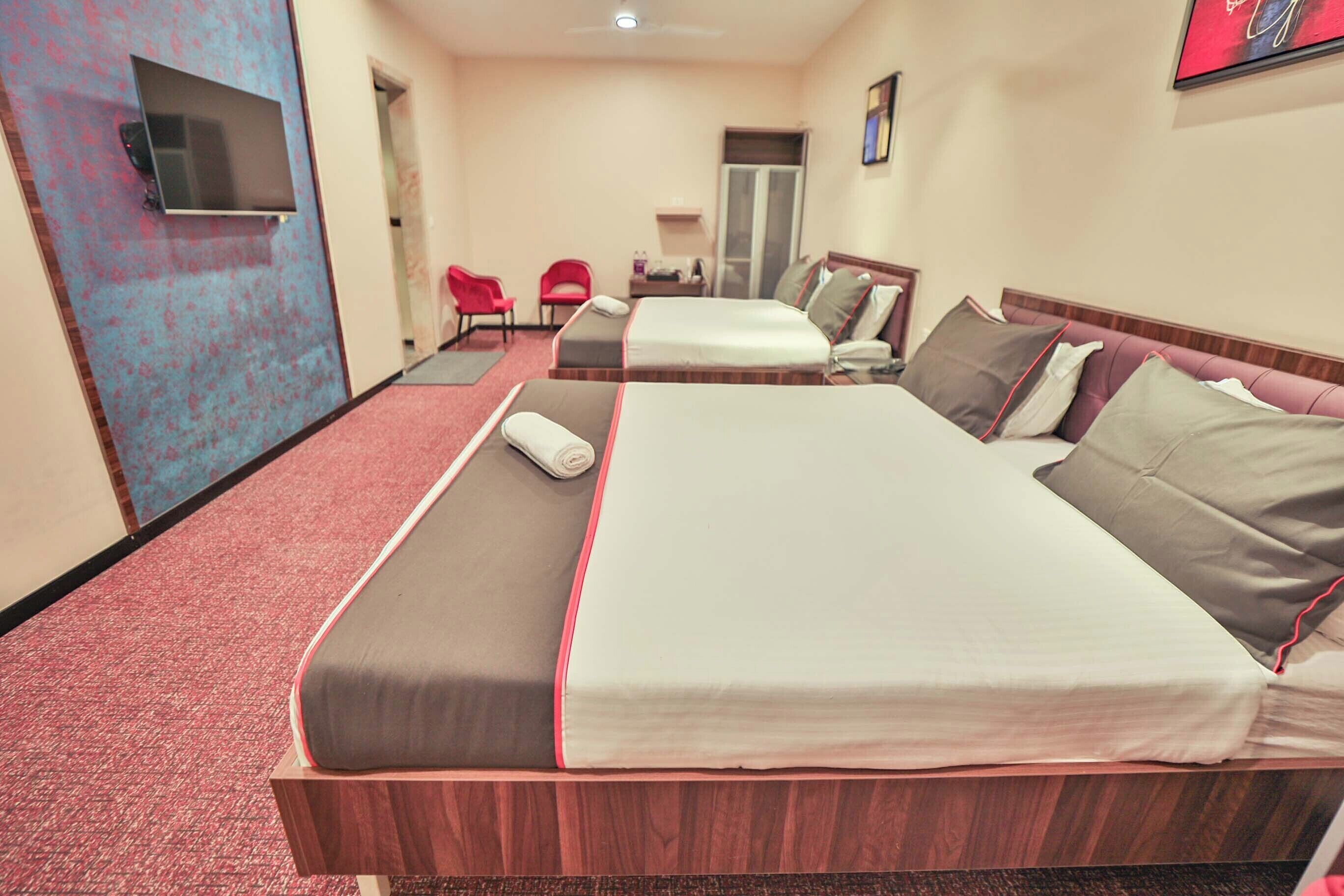 A hotel room with two beds, a TV mounted on the wall, a red chair, and a door leading to another room.