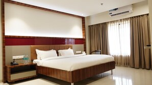 A hotel room with a large bed, a nightstand with a phone and water bottles, a wall-mounted air conditioner, and a window with curtains.