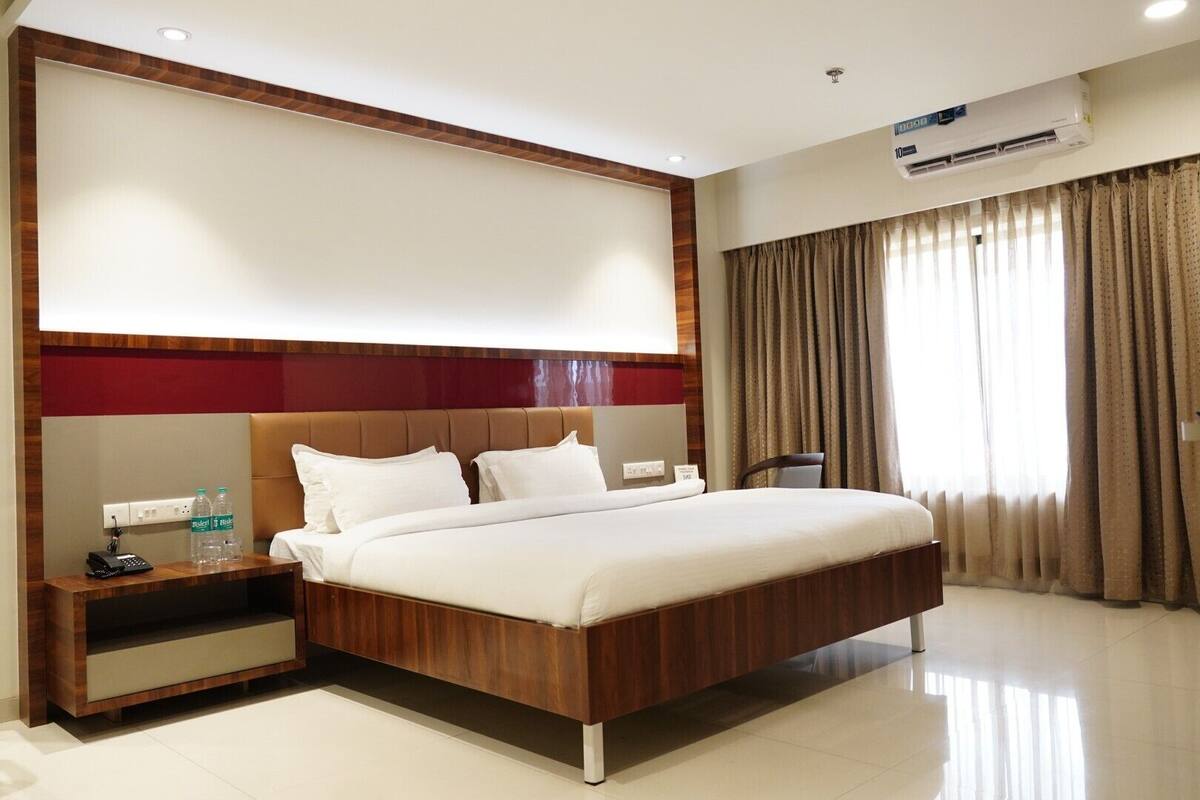 A hotel room with a large bed, a nightstand with a phone and water bottles, a wall-mounted air conditioner, and a window with curtains.