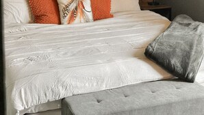 A bedroom with a tufted headboard, a bed with white bedding and orange and gray pillows, a gray bench, a nightstand with a lamp, and a wall with a fern-patterned wallpaper.