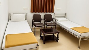 Une petite chambre propre, avec deux lits simples, une petite table, deux chaises et une fenêtre avec des rideaux marron.