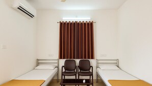 Une chambre avec deux lits simples, un ventilateur de plafond, un appareil de climatisation, une fenêtre avec des rideaux et une petite table avec des chaises.