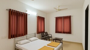Une chambre avec un lit simple, un cadre en métal, deux chaises, une petite table et deux ventilateurs de plafond.