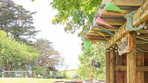 A rustic wooden building with a thatched roof, surrounded by a fenced area with a small boat and a flag. - Stardust (Ventnor)