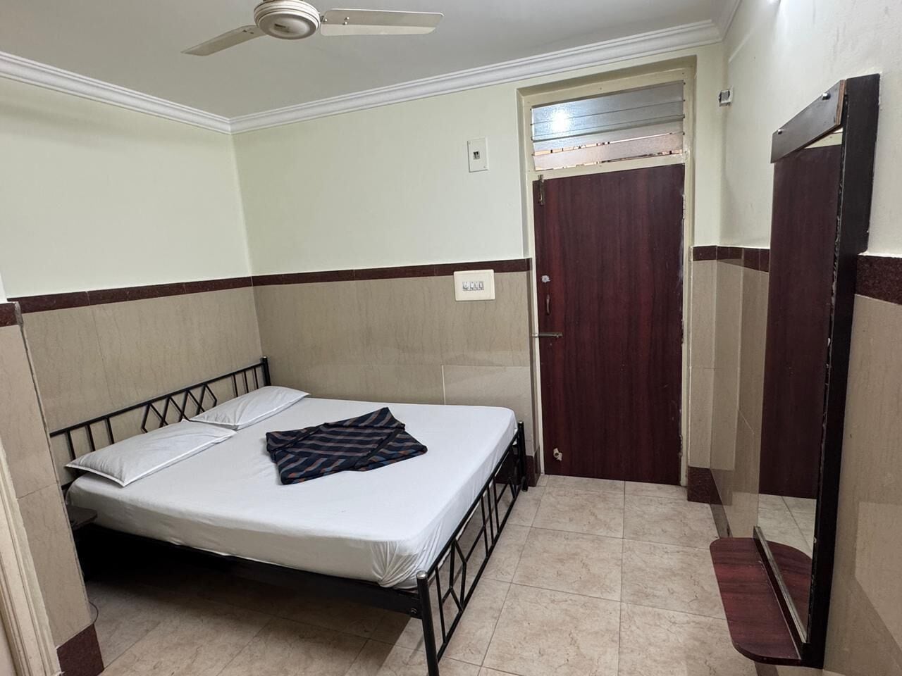 Un lit simple avec un matelas blanc et une couverture à rayures, un ventilateur de plafond, une porte en bois et un sol carrelé.