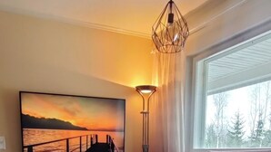 Olohuoneessa on sohva, penkki, seinälle asennettu televisio ja lattiavalaisin.