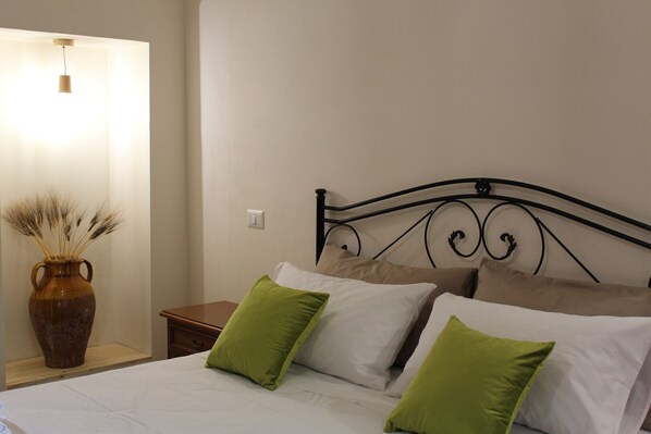 Una cama bien hecha con almohadas blancas y verdes, un jarrón marrón con plantas secas y una mesita de noche de madera.