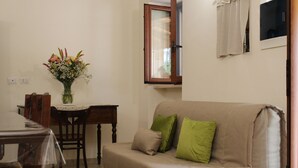 Una sala de estar con un sofá color beige, dos cojines verdes, una mesa de comedor de madera con sillas y un florero con flores.