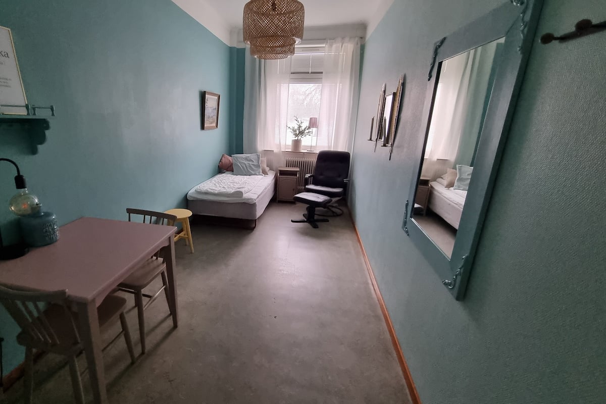 A compact room with a bed, a desk, and a chair. There is a shelf with a sign on the wall and a hanging light fixture.