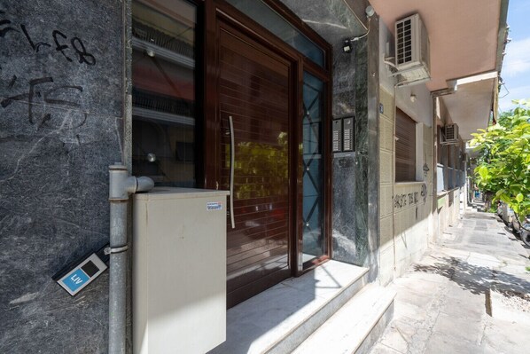 Une rue étroite, bordée d’un immeuble qui se distingue par une grande porte en bois et un petit réfrigérateur blanc placé à l’extérieur.