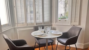 Un coin repas avec une table ronde, deux chaises et une vue dégagée grâce à de grandes fenêtres équipées de volets.
