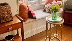 Une pièce avec une grande fenêtre en saillie, un banc en bois, une petite table ornée d’un vase de fleurs et un téléviseur fixé au mur.