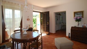 Un coin repas avec une table ronde, des chaises, un lustre et une vue sur la cuisine et le jardin, grâce à une porte ouverte.