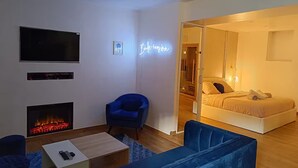 Moderni hotellihuone, jossa on taulutelevisio, takka, sininen nojatuoli, sohva ja sänky, jonka yläpuolella on neonvalomainos.