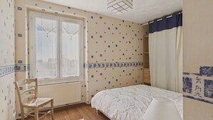 A bedroom with a bed, a chair, a nightstand with a lamp, a window with curtains, and patterned wallpaper. - The port and beaches nearby - 2 adults 2 children (Concarneau)