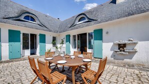 Une cour avec une table et des chaises en bois rondes, un sol en pierre et un bâtiment blanc avec des volets bleus et une cheminée en pierre.