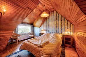 Cozy attic bedroom with a large bed, wooden nightstand, and wicker chair, set against a backdrop of floral wallpaper and wooden paneling.