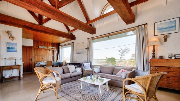 Un salon spacieux avec un plafond haut, des poutres apparentes, une grande fenêtre avec des rideaux, un ensemble de canapés, une table basse, des fauteuils en osier et un meuble en bois.
