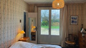 Une chambre d’hôtel avec un lit, une commode en bois, une chaise, un bureau avec une lampe et une grande fenêtre donnant sur un jardin.