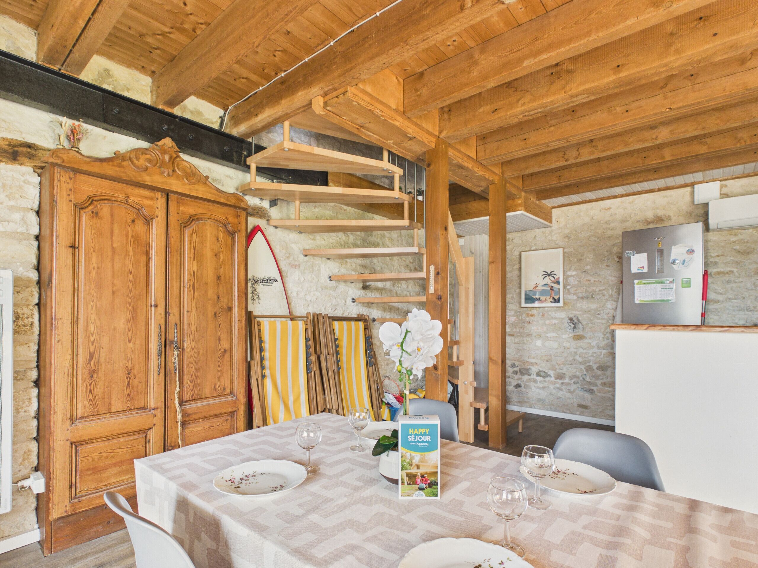 Un coin repas avec une table dressée pour deux personnes, un buffet en bois, une planche de surf et un réfrigérateur.