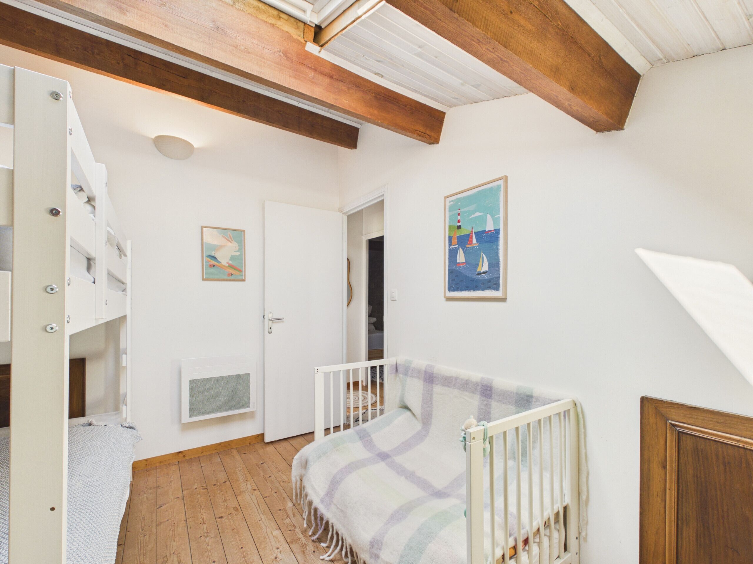 Une pièce avec un plafond en bois, une porte blanche, un berceau avec une couverture, une petite fenêtre et un tableau accroché au mur.