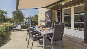 Eine Terrasse im Freien mit Tisch und Stühlen, einem großen Sonnenschirm und Blick auf Garten und Gebäudefassade.