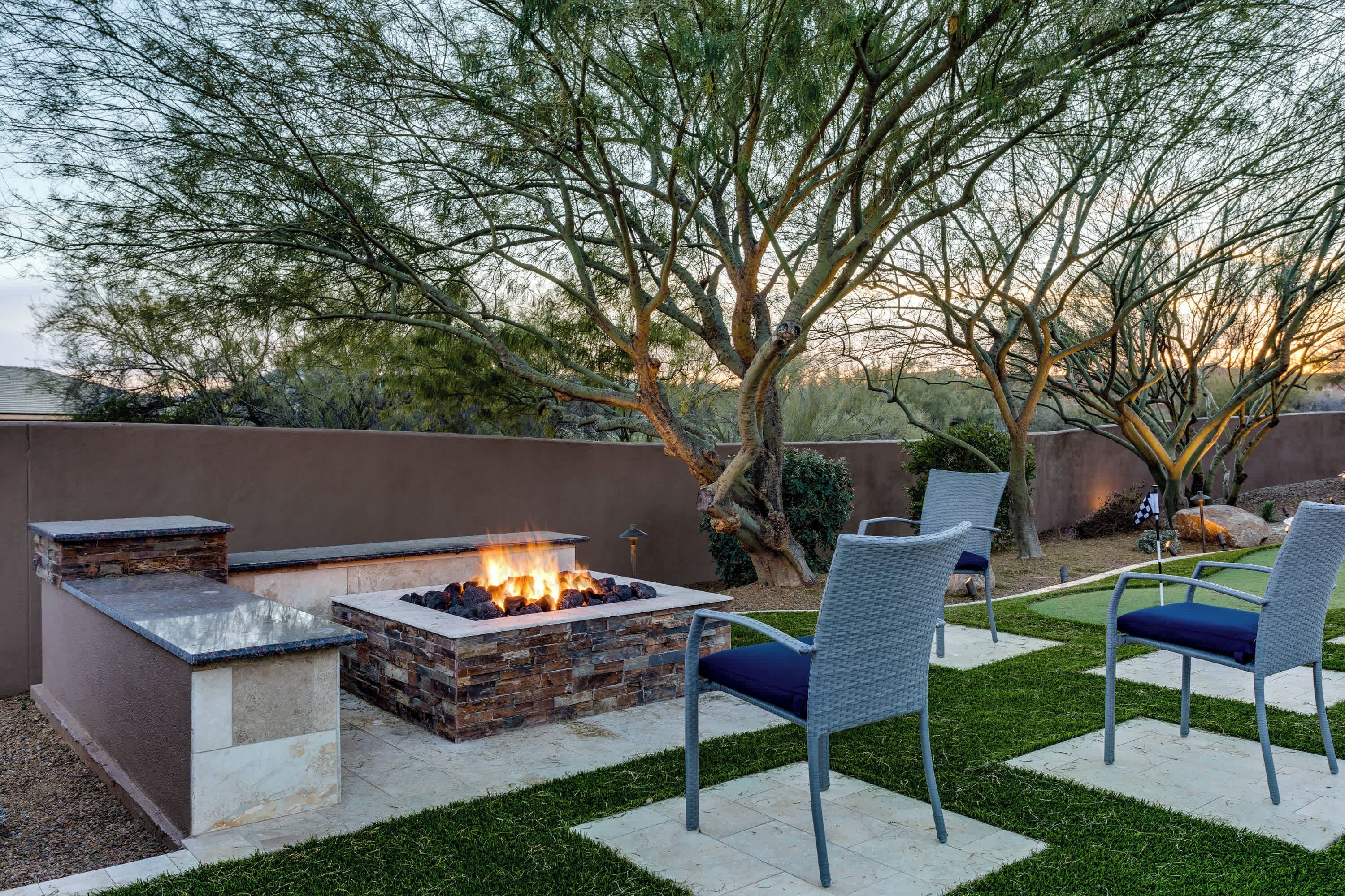An outdoor patio with a stone fire pit, two wicker chairs, and a well-maintained lawn.