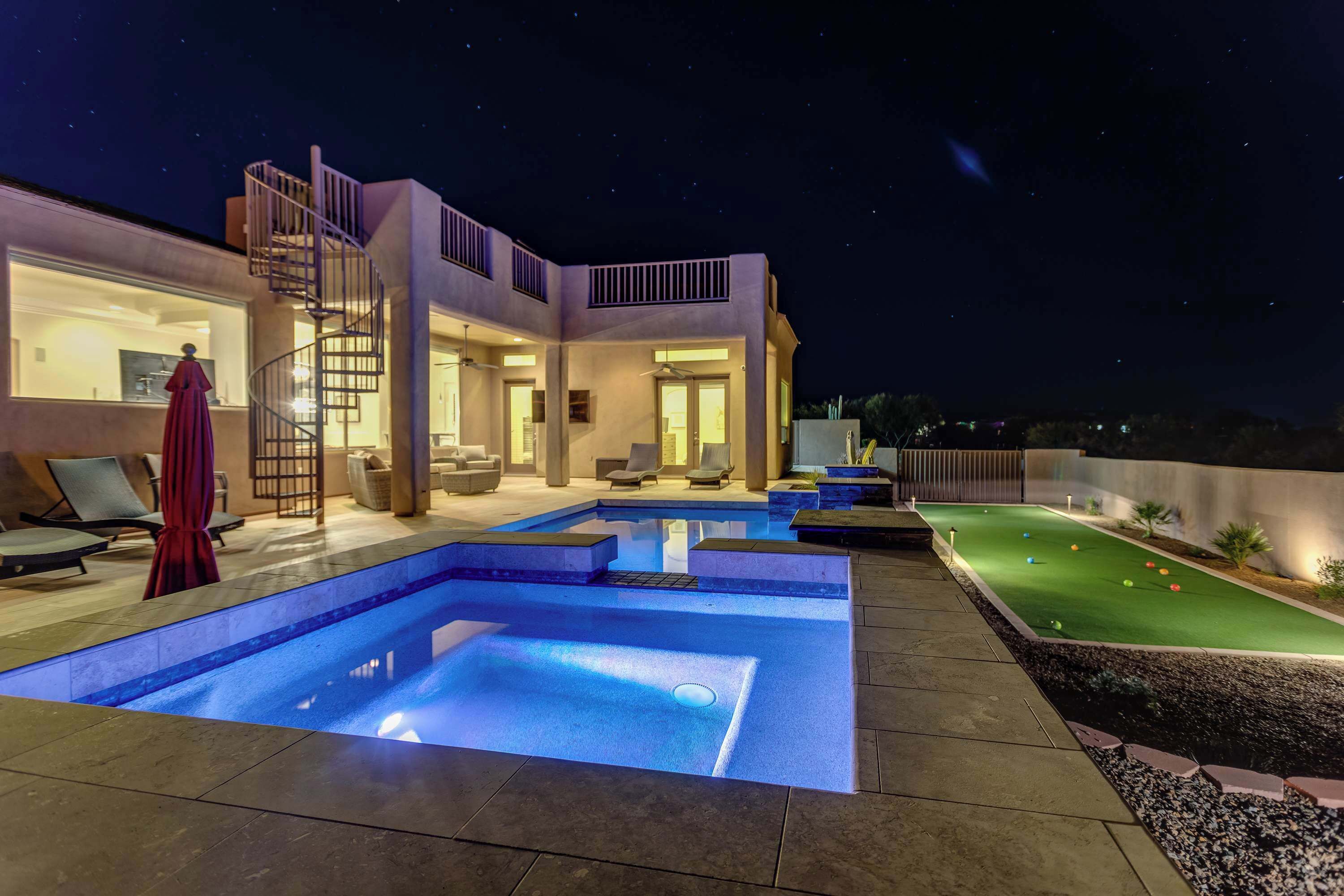 A modern outdoor pool area with a well-lit deck, lounge chairs, and a small putting green.