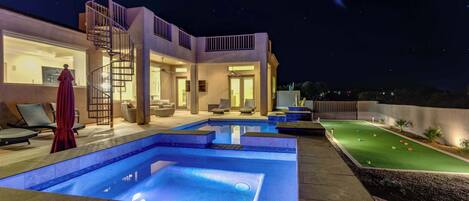 A modern outdoor pool area with a well-lit deck, lounge chairs, and a small putting green.