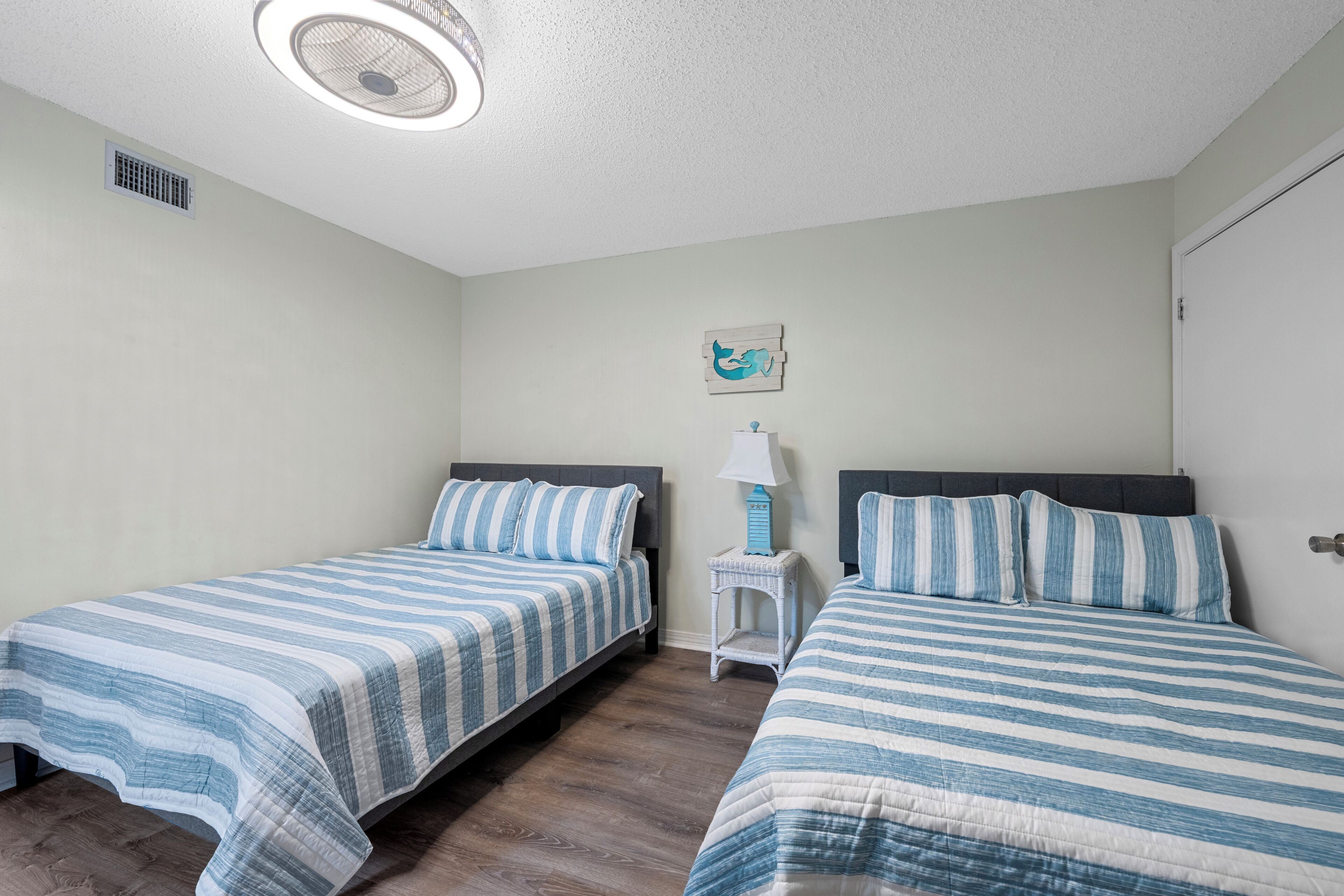 A bedroom with two beds, a ceiling fan, a small table with a lamp, and a wall-mounted picture.