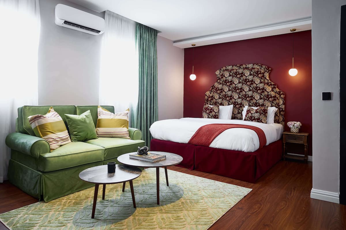 A hotel room with a green sofa, a round coffee table, a large bed with a red headboard, and a small bedside table.