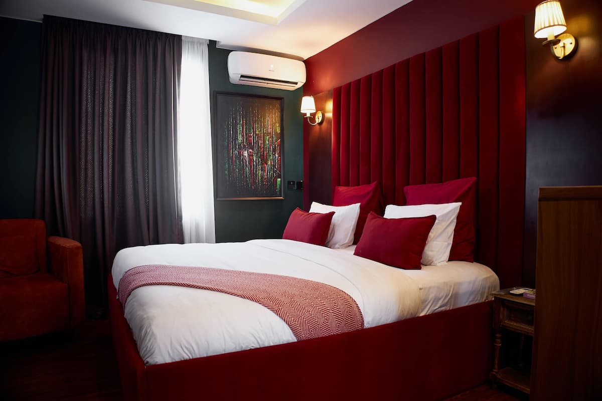A hotel room with a large bed, red and white bedding, a red armchair, and a wall-mounted air conditioner.