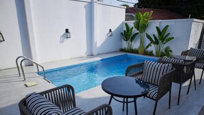 A swimming pool with a white wall, surrounded by wicker furniture and a table. - The Hendon House (Lagos)