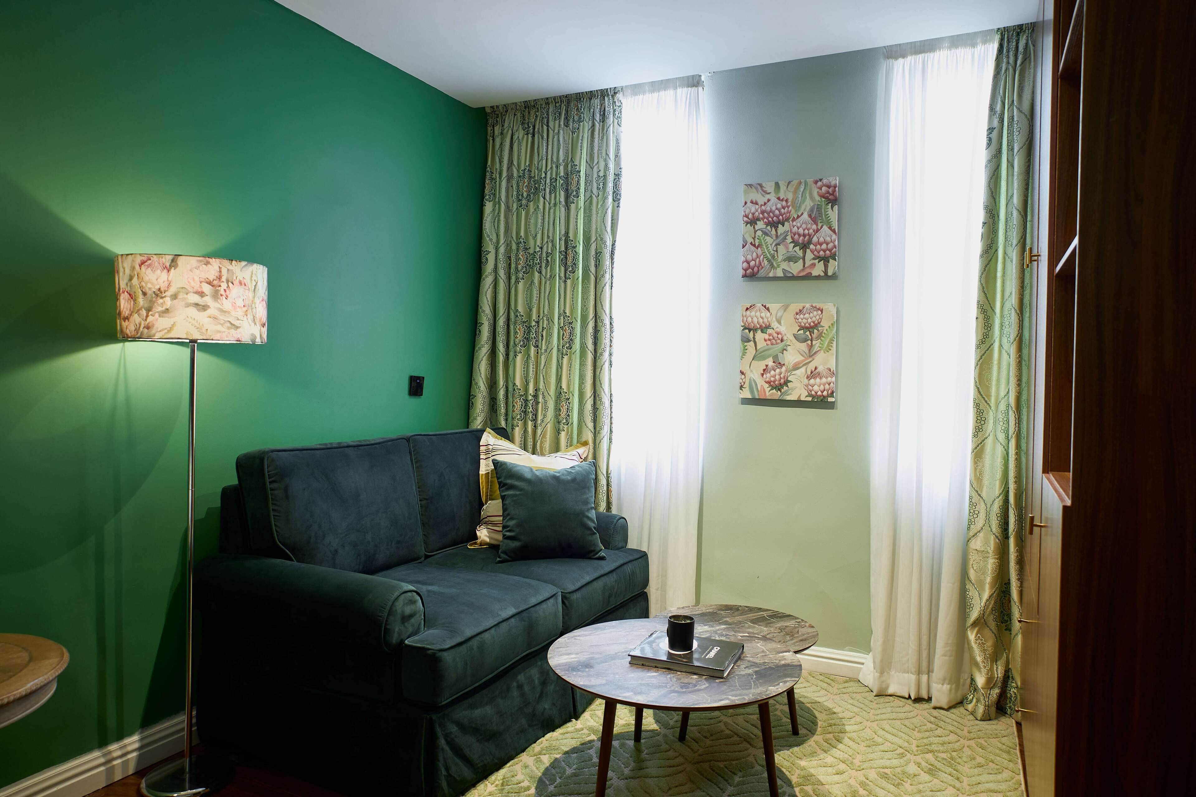 A living room with a green wall, a dark green sofa, a round coffee table with a book and a cup, a floor lamp, and floral curtains.