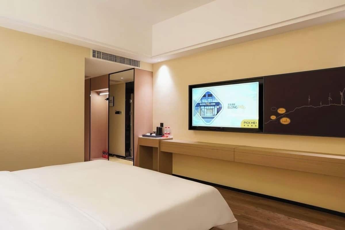 A hotel room with a bed, a TV mounted on the wall, and a small table with a bottle and a cup.