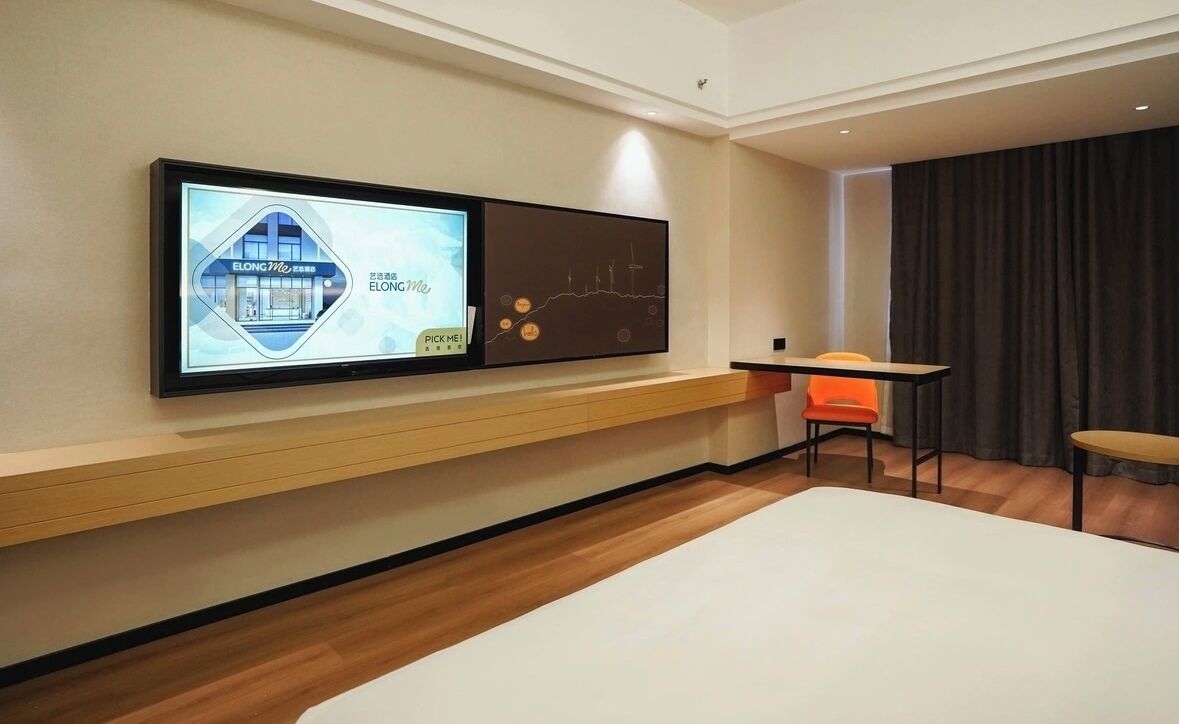 A hotel room with a large flat-screen TV mounted on the wall, a wooden floor, a bed, a small table, and an orange chair.