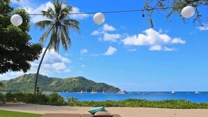 A beach view with a lounge area, palm trees, and a clear sky. - Elegant Two-Bedroom Condo with Oversized Terrace, Resort Pools & Beach Club Access (Playas del Coco)