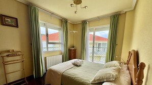 A hotel room with a bed, a wooden headboard, a bedside table, a lamp, a radiator, a window with curtains, and a view of buildings outside. - Comfortable apartment near the beach (Isla)