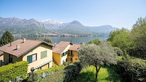 A lakeside property with a mountain backdrop, featuring a yellow building with a tiled roof and a green window.