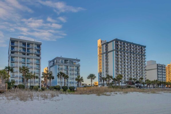 Un hotel frente a la playa con edificios de varios pisos, balcones y palmeras.