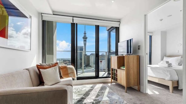 A modern hotel room with a large window, a sofa, a bed, a TV, and a wooden cabinet.