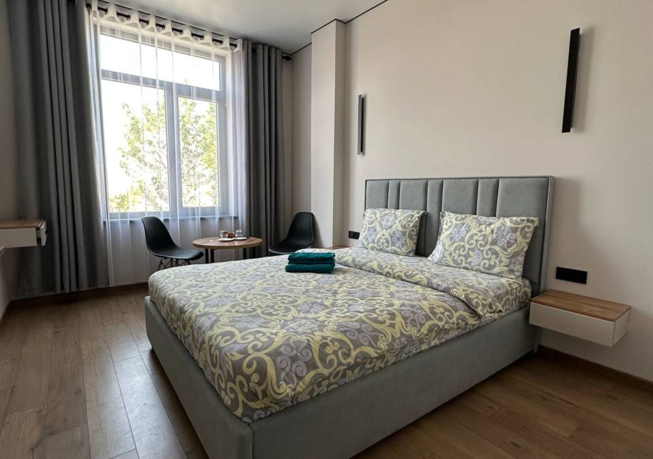 A modern hotel room with a large bed, a small desk, a chair, and a window with curtains.