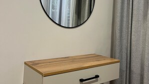 A white bedside table with a wooden top and a black drawer handle, positioned next to a grey curtain and a round mirror on the wall. - Tikhii Zamok (Bishkek)