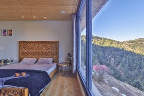 A bedroom with a large bed, a wooden headboard, a small table, and a view of a forested hillside.