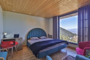 A bedroom with a bed, a desk, and a chair. There is a window with a view of a mountain. - Suhag Valley View (Dalhousie)