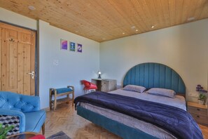 A bedroom with a wooden ceiling, a bed with a blue headboard, a blue sofa, a wooden chair, and a small table with a lamp. - Suhag Valley View (Dalhousie)